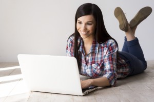 Spending her free time in the net. Attractive young woman working on laptop and smiling while lying on the hardwood floor